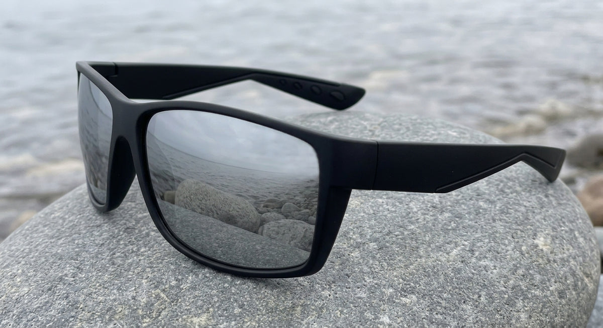 Lunettes polarisantes à miroir argent et monture noire mate posées sur un gros galet lisse au bord de l'eau