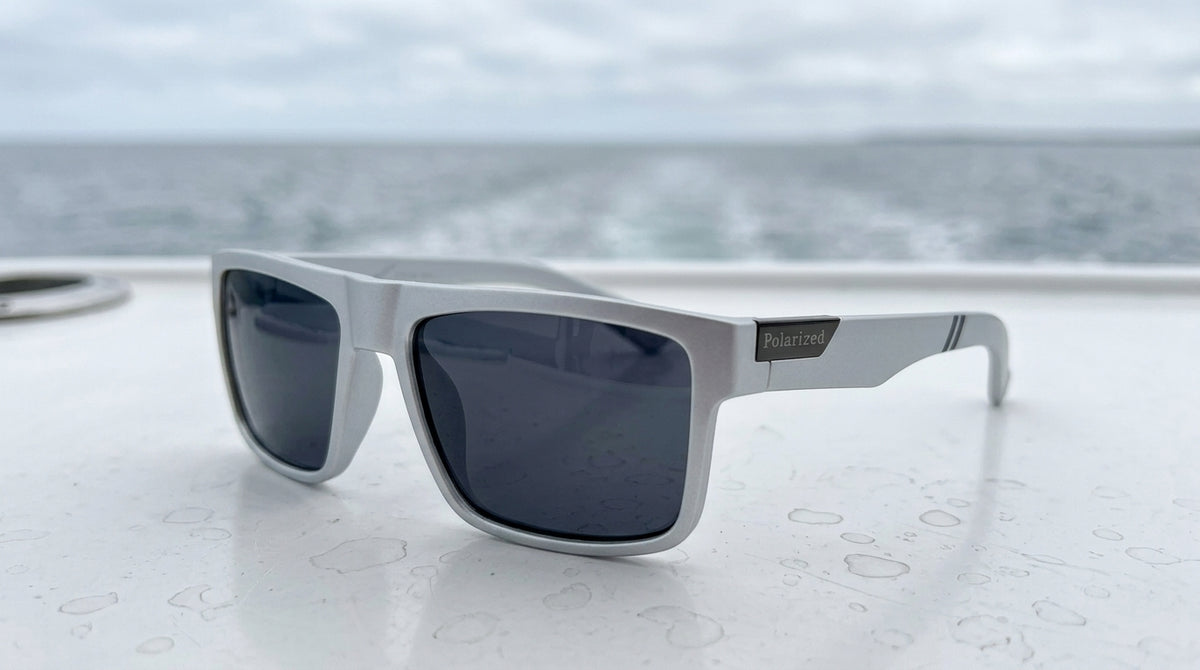 Lunettes polarisantes gris fumé à monture grise posées sur un pont de bateau blanc perlé de gouttes, sillage en mer derrière