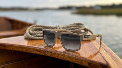 Lunettes polarisantes gris fumé à monture kaki posées sur le plat-bord en bois verni d'une barque, un cordage lové derrière, estuaire en arrière-plan