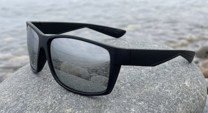 Lunettes polarisantes à miroir argent et monture noire mate posées sur un gros galet lisse au bord de l'eau