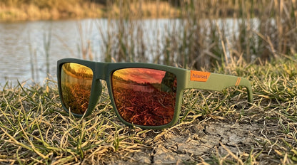 Lunettes polarisantes à miroir rouge et monture vert foncé posées sur une berge d'herbe rase au bord d'un étang, roseaux en arrière-plan