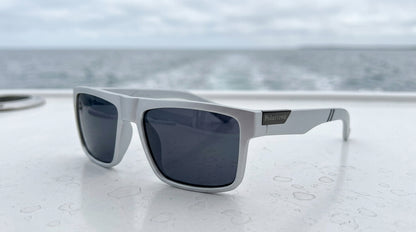 Lunettes polarisantes gris fumé à monture grise posées sur un pont de bateau blanc perlé de gouttes, sillage en mer derrière