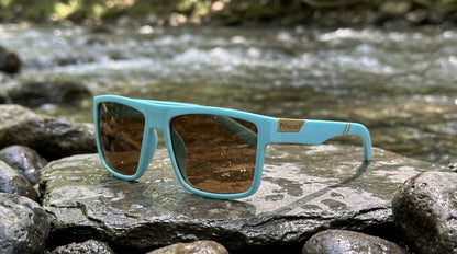 Lunettes polarisantes teinte ambre à monture turquoise posées sur une pierre mouillée au bord d'une rivière, eau courante en arrière-plan