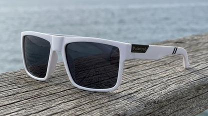 Lunettes polarisantes gris fumé à monture blanche posées sur une planche de bois flotté, mer en arrière-plan